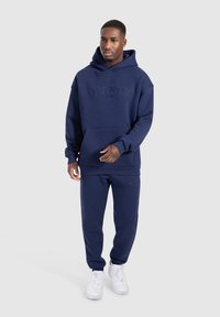 Felpa con cappuccio di colore blu navy e pantaloni abbinati in tessuto morbido, con tasca frontale e dettagli del logo sul petto e sulla gamba.