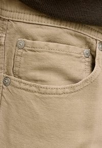 Poche de pantalon en tissu beige avec rivets métalliques estampillés "JACK & JONES" et surpiqûres doubles visibles sur un matériau texturé.