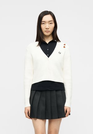Jeune femme portant un cardigan court blanc avec des écussons en forme de cœur orange sur une chemise noire à col, et une jupe plissée foncée, debout devant un fond blanc.