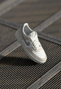 Nike Air Force 1 toss, mis on valge ja helebeeži värvigammaga, nahast ja seemisnahast materjalid, perforatsiooniga varvastele ning lamedad paelad.