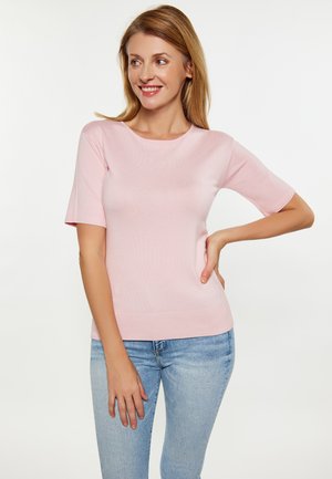 Kvinde smiler og kigger mod højre iført en lyserød kortærmet top og lyseblå jeans mod en ensfarvet hvid baggrund.