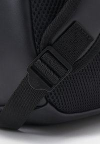 Black bag detail featuring smooth synthetic material, a textured mesh section, and a black plastic buckle. Strap has an adjustable design.