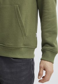 Sudadera verde oliva con un bolsillo tipo canguro, puños y dobladillo acanalados, textura suave y una pequeña etiqueta de la marca en la costura lateral. Combinada con jeans oscuros.