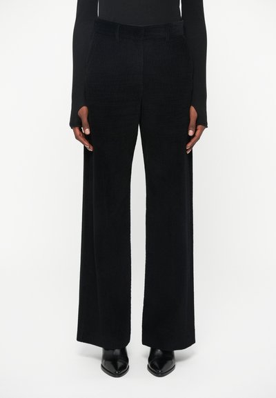Pantaloni in corduroy neri a gamba larga con texture a righe verticali, dotati di tasche frontali e una vita liscia, abbinati a scarpe nere a punta.