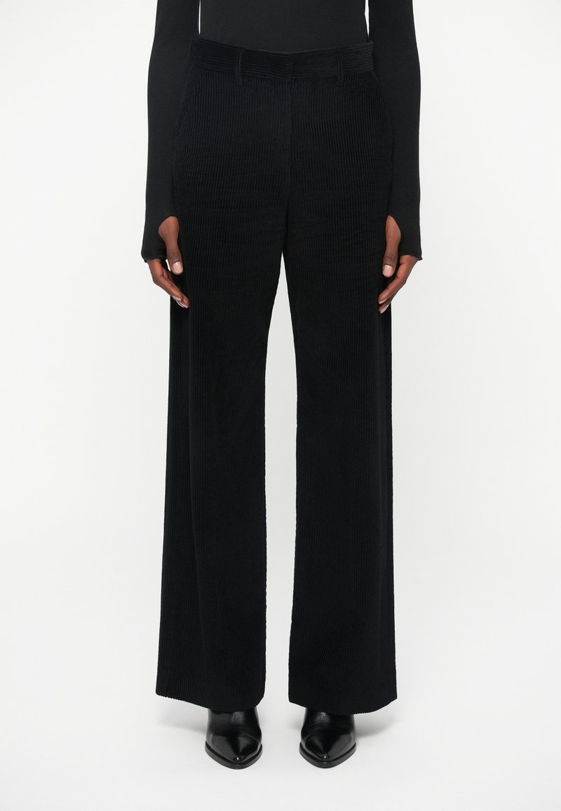 Black wide-leg corduroy trousers with vertical ribbed texture, featuring front pockets and a smooth waistband, paired with black pointed shoes.