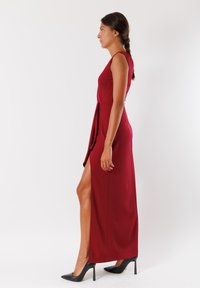 Abito senza maniche color bordeaux con uno spacco laterale, realizzato in tessuto morbido. Presenta un corpetto aderente e arriva fino a terra. Tacchi alti neri.