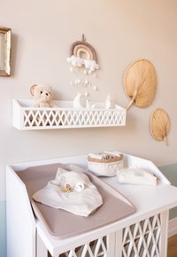 Stație de schimbat scutece în tonuri neutre, cu un ursuleț de pluș, suzete, jucării moi, coșuri de depozitare, un carusel decorativ în formă de curcubeu și frunze de palmier montate pe perete.