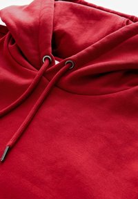 Felpa rossa con finitura testurizzata, dotata di cappuccio regolabile con cordoncino e occhielli metallici. Il design è semplice e pulito, concentrandosi su un colore uniforme.