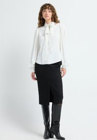 Hvid bluse med en stor sløjfe, puffede ærmer og knaplukning, parret med en sort knælang nederdel og blanke sorte knæhøje støvler.