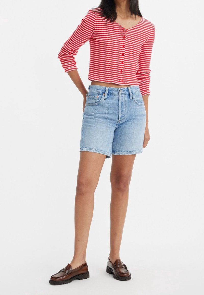 Femme portant un cardigan court rayé rouge et blanc, un short en denim bleu clair et des mocassins marron, debout avec une main sur la hanche.