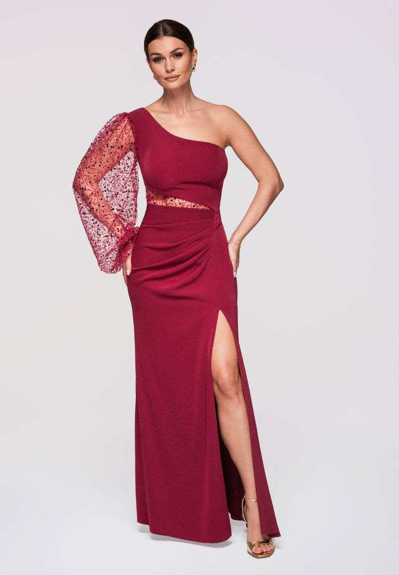 Femme portant une robe bordeaux asymétrique avec une manche longue en tulle ornée de détails et une fente haute sur la cuisse, posant avec une main sur la hanche.
