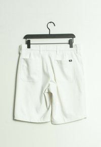 Pantalones cortos blancos hechos de una tela suave, con dos bolsillos traseros y un pequeño logo en el bolsillo derecho. Colgados de una percha negra con clip.