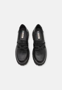 Mocasines de cuero negro con una textura suave, que presentan un acento de cadena distintivo y suelas de goma gruesas para mayor agarre.