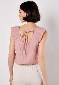 Top rosa chiaro con un tessuto testurizzato e stropicciato. Presenta maniche a volant e una schiena bassa con chiusura a nodo. Abbinato a pantaloni di colore chiaro.