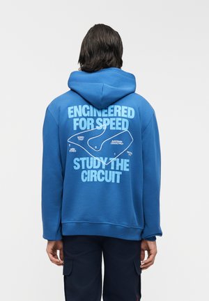 Άτομο που φοράει μπλε φούτερ με κουκούλα, με χάρτη αγωνιστικής πίστας και κείμενο "Σχεδιασμένο για ταχύτητα" και "Μελέτη του κυκλώματος" στην πλάτη.