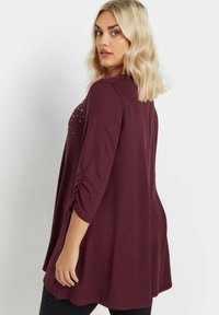 Burgundy tunika yläosa, jossa on kolme neljännesosa hihaa, kerrottuja yksityiskohtia, pehmeää kangasta ja koristeellisia niittejä yhdessä olkapäässä. Leveä leikkaus.