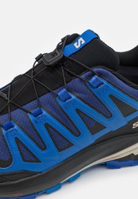 Blå och svarta träningsskor med en texturerad tygöverkonstruktion, dynamiska designlinjer och ett säkert snörningssystem med silikonreglage.