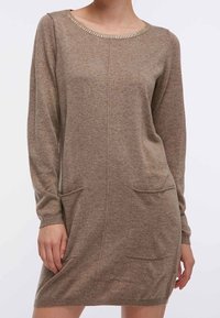 Femme portant une robe tricotée à manches longues, de couleur marron clair, avec deux poches à l'avant et un col rond orné de petites perles.