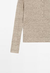 Haut en tricot côtelé beige à manches longues avec une patte de boutonnage comportant cinq petits boutons sur fond blanc.