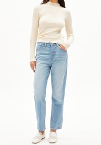 Cremfarbene, gerippte Langarmbluse, kombiniert mit hellblauen, weit geschnittenen Denim-Jeans. Das Model trägt weiße, geflochtene Slipper.