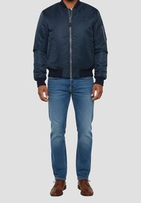 Homme portant une veste bomber bleu marine, un jean bleu et des chaussures en cuir marron à lacets, debout devant un fond gris.