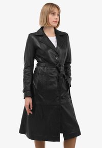 Zwarte leren trenchcoat met een tailleband, grote revers en knopen aan de voorkant. Beschikt over twee zijzakken en een lengte tot halverwege het kalf. Gladde textuur.