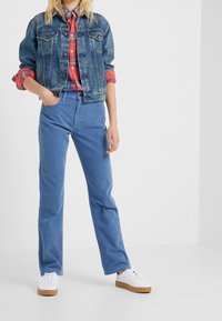 Personne portant une veste en denim par-dessus une chemise à carreaux rouge, un pantalon en velours côtelé bleu et des baskets blanches, se tenant devant un fond blanc uni.
