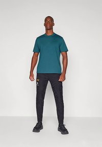 Man med en teal kortärmad skjorta, svarta UFC-märkta joggingbyxor och svarta sneakers, stående mot en ljus enfärgad bakgrund.
