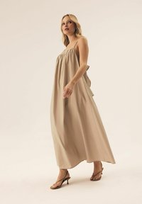 Beżowa sukienka maxi z cienkimi ramiączkami, marszczonym korpusem, zwiewnym materiałem oraz falbaniastymi dodatkami po bokach. Połączona z wąskimi sandałkami na obcasie.