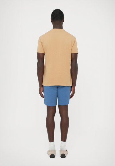 Világosbarna pamut póló rövid ujjakkal, kék rövidnadrággal párosítva. A modell hátulról látható, sima textúrákkal és egyszerű dizájnnal.
