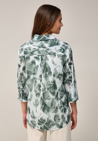 Grön och vit mönstrad blus med krage, långa uppvikta ärmar och rundad nederkant. Tyget har ett Struktur och abstrakt mönster.