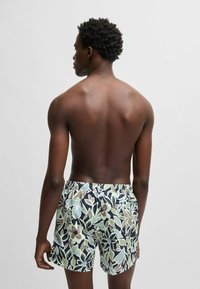 Simshorts i svart med ett blommönster som har gröna blad och ljusa blomsteraccenter. Elastisk midja, lös passform, medellängd.