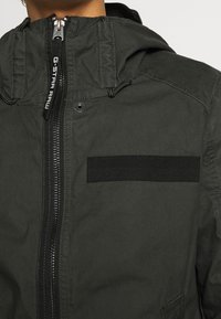 Mörkgrön huva-jacka i lätt material. Har en frontdragkedja, en ficka med lappar och justerbara snören på huvan.