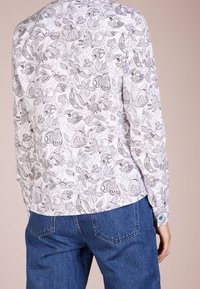 Langärmliges Hemd aus weißem Stoff mit aufwendigen schwarzen Blumen- und Schmetterlingsmustern, mit Knopfmanschetten und abgerundetem Saum.
