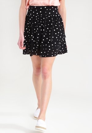 Femme marchant portant une jupe noire à couches avec des pois blancs, un haut rose pâle et des chaussures blanches slip-on avec des semelles tressées.