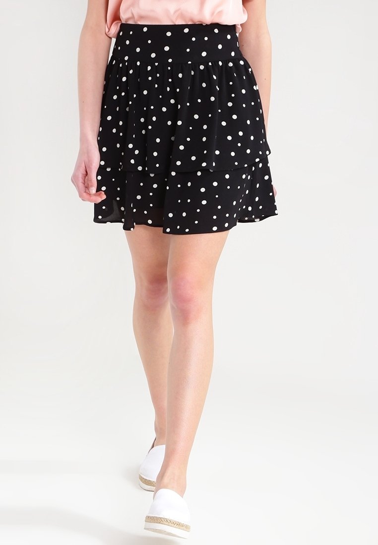 Femme marchant portant une jupe noire à couches avec des pois blancs, un haut rose pâle et des chaussures blanches slip-on avec des semelles tressées.