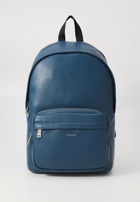 Zaino in pelle blu con forma arrotondata, texture liscia, tasca frontale con zip e dettaglio con logo. Dotato di manico superiore nero e spallacci regolabili.