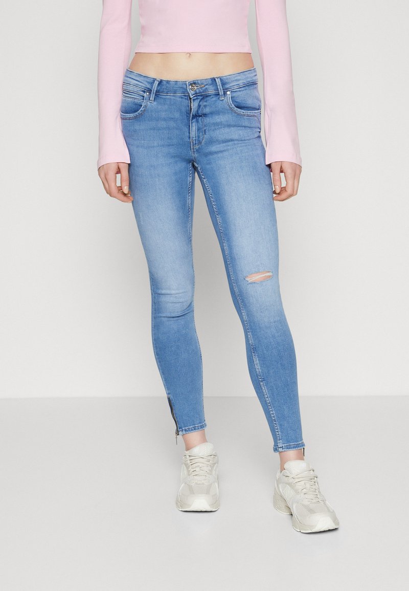 Λεπτά μπλε τζιν με μεσαίου ύψους μέση, ανοιχτό πλύσιμο και φθαρμένο γόνατο. Συνδυασμένα με μια παλ-ροζ μπλούζα μακριού μανικιού και ουδέτερα αθλητικά παπούτσια.