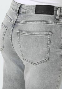 Grey denim jeans featuring a medium rise, back pockets, and a black leather label on the waistband. Textured fabric with visible stitching.