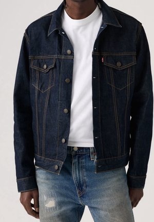 Mand iført mørkeblå denimjakke over hvid T-shirt og lyseblå slidte jeans, stående foran ensfarvet baggrund.