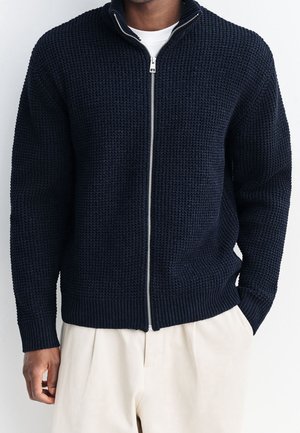 Homme portant un cardigan zippé bleu marine texturé par-dessus une chemise blanche et un pantalon beige, debout avec une main dans la poche.