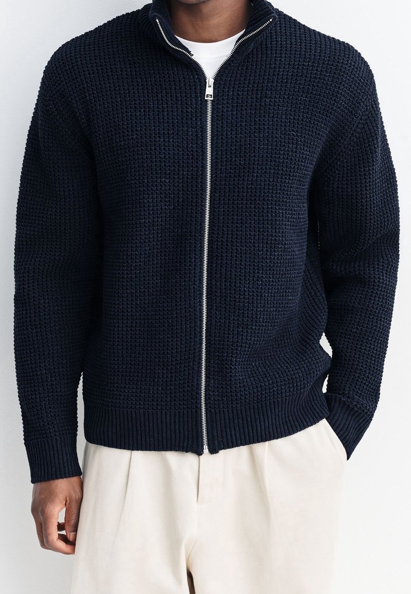 Homme portant un cardigan zippé bleu marine texturé par-dessus une chemise blanche et un pantalon beige, debout avec une main dans la poche.