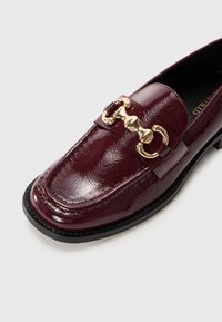 Mocassin en cuir verni bourgogne avec un ornement en chaîne dorée, bout rond et une semelle noire basse, avec une finition texturée.