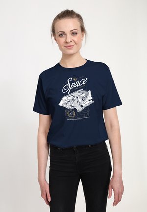 Kobieta nosząca granatowy t-shirt z białą grafiką statku kosmicznego i słowem „Space” pisanym kursywą na przodzie, w połączeniu z czarnymi spodniami.