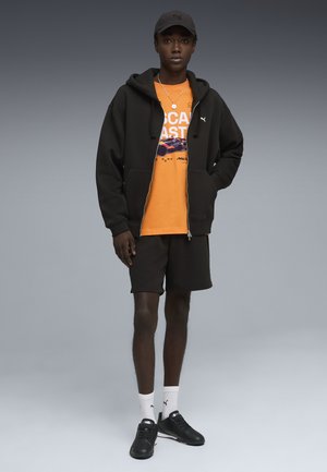 Jeune homme portant une casquette noire Puma, un sweat à capuche zippé noir, un t-shirt graphique orange, un short noir, des chaussettes blanches Puma et des baskets noires.