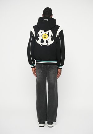 Melns kapucis ar baltām akcentiem un lielu grafiku, kas attēlo dzeltenu smaidošu seju mugurpusē. Apvienots ar vaļīgām pelēkām džinsām un sporta apaviem.