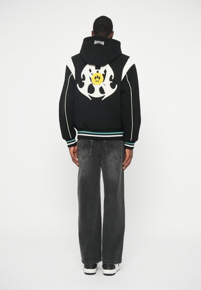 Schwarzer Hoodie mit weißen Akzenten und einem großen grafischen Motiv, das ein gelbes lächelndes Gesicht auf dem Rücken zeigt. Kombiniert mit lockeren grauen Jeans und Sneakern.