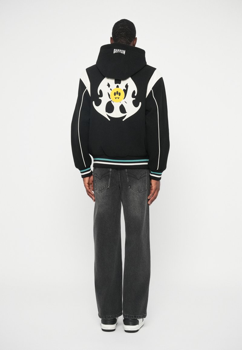 Schwarzer Hoodie mit weißen Akzenten und einem großen grafischen Motiv, das ein gelbes lächelndes Gesicht auf dem Rücken zeigt. Kombiniert mit lockeren grauen Jeans und Sneakern.