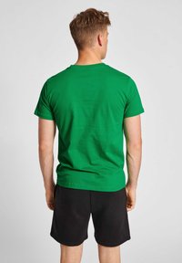 Grön bomullströja med korta ärmar och rund halsringning, bärs med svarta shorts, sedd bakifrån för att visa en slät yta.
