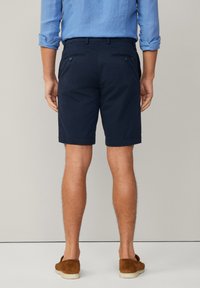 Shorts en coton bleu marine avec une coupe ajustée, dotés de poches latérales et de deux poches arrière avec fermetures à bouton. Portés avec des chaussures à enfiler marron.